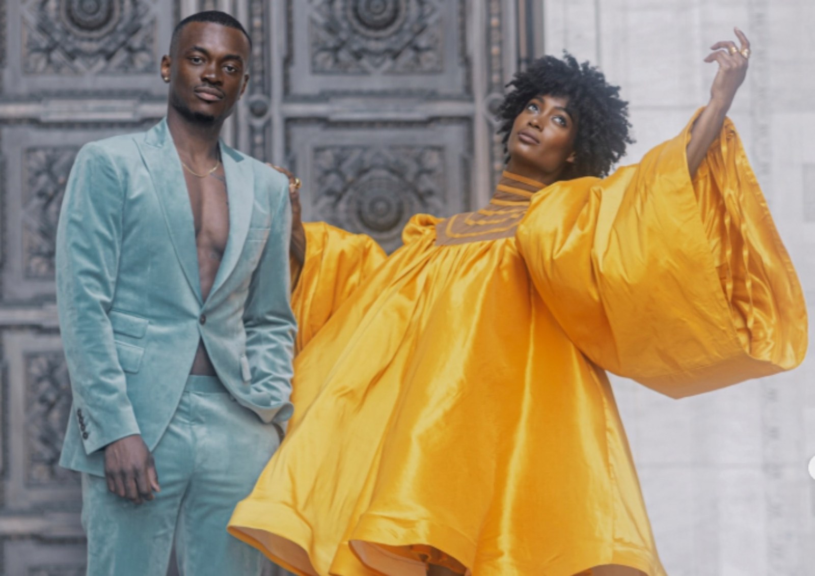 Two models on stone steps before ornate bronze doors, one in a pale green velvet suit and one in a wide-sleeved marigold silk gown, both by Chuks Collins