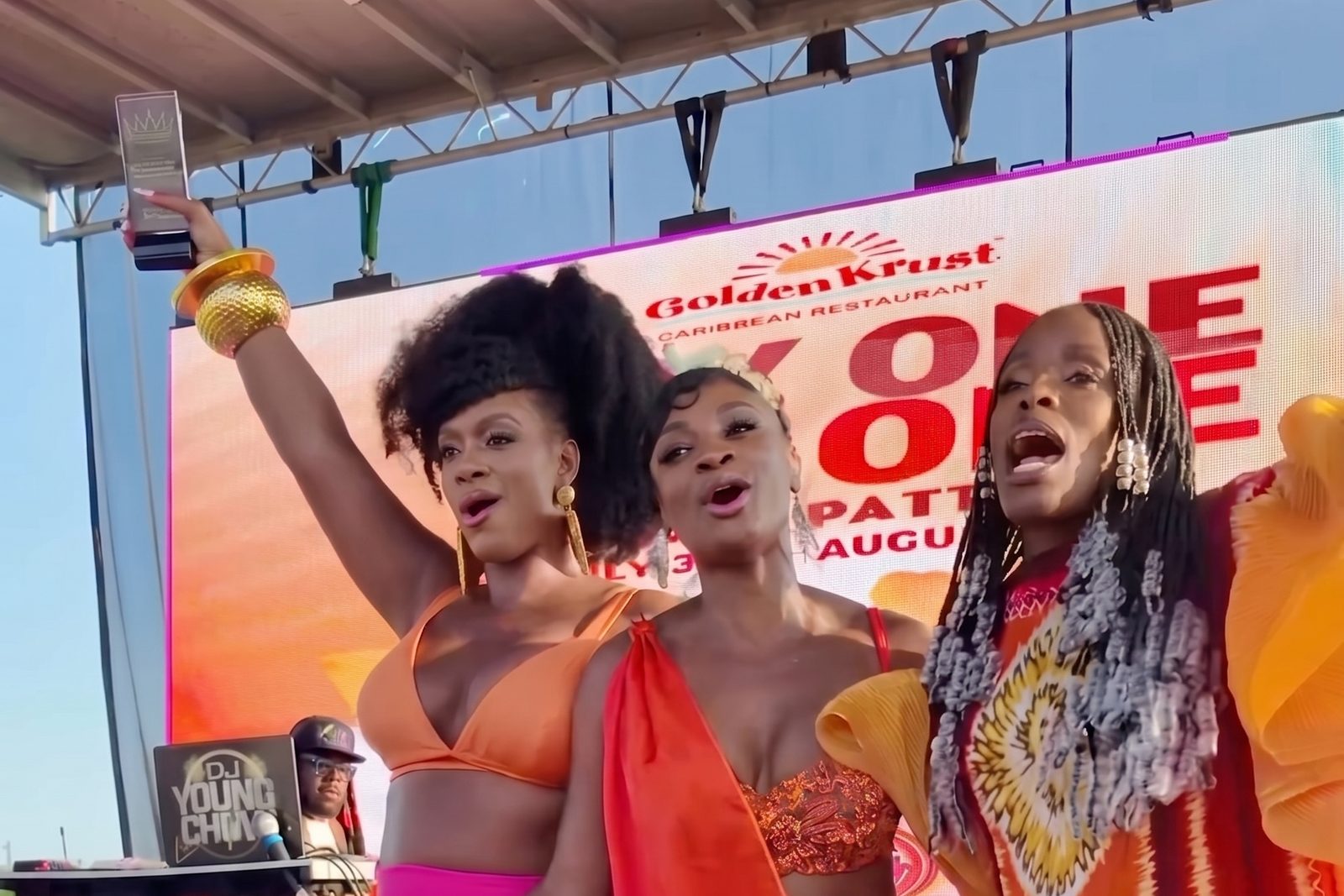 Three performers on the CurlFest stage holding an award, in front of a screen carrying Golden Krust Caribbean Restaurant branding