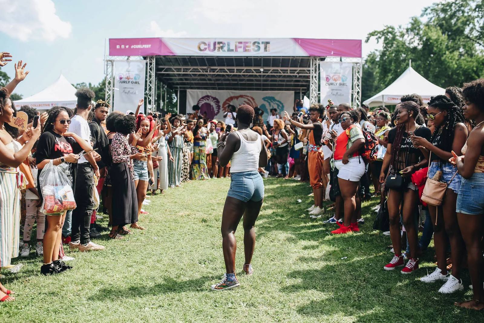 A CurlFest crowd lining both sides of a grass runway in front of the main stage, with Curly Girl Collective banners either side