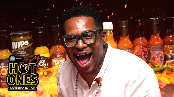 A guest in green-framed glasses laughing in front of a row of hot sauces, with the Hot Ones Caribbean Edition logo