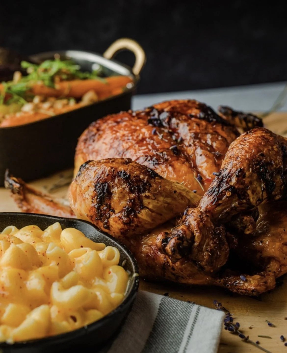 A glazed whole rotisserie chicken beside a skillet of macaroni and cheese and a dish of roasted carrots