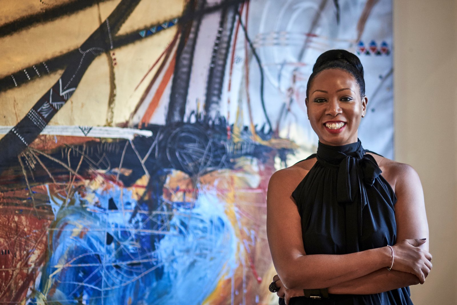 A smiling woman in a black sleeveless top with a tied collar, arms folded, in front of a large abstract painting
