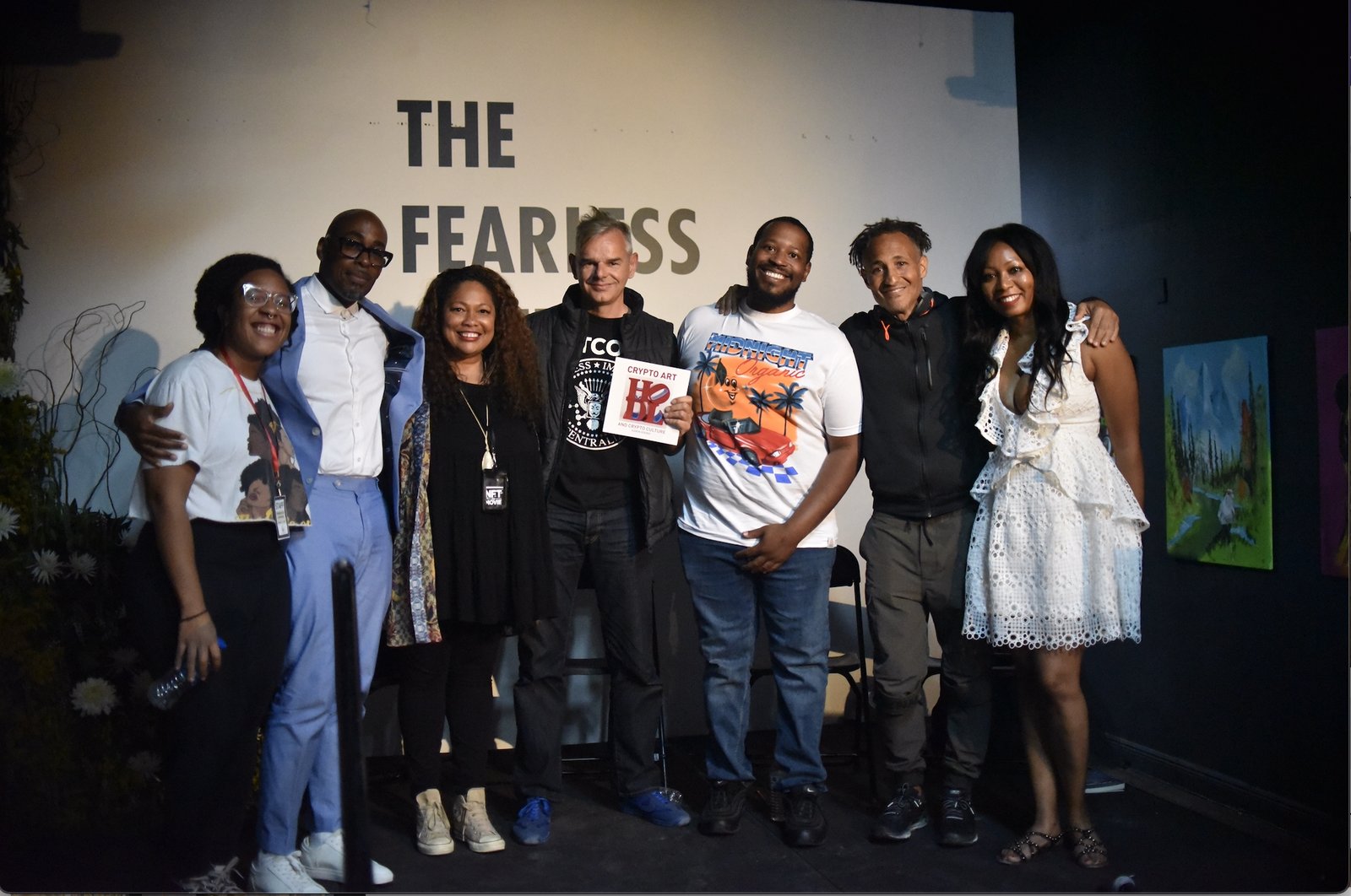 Seven people standing together on a stage in front of a wall painted with The Fearless Artist name, one holding a book titled Crypto Art