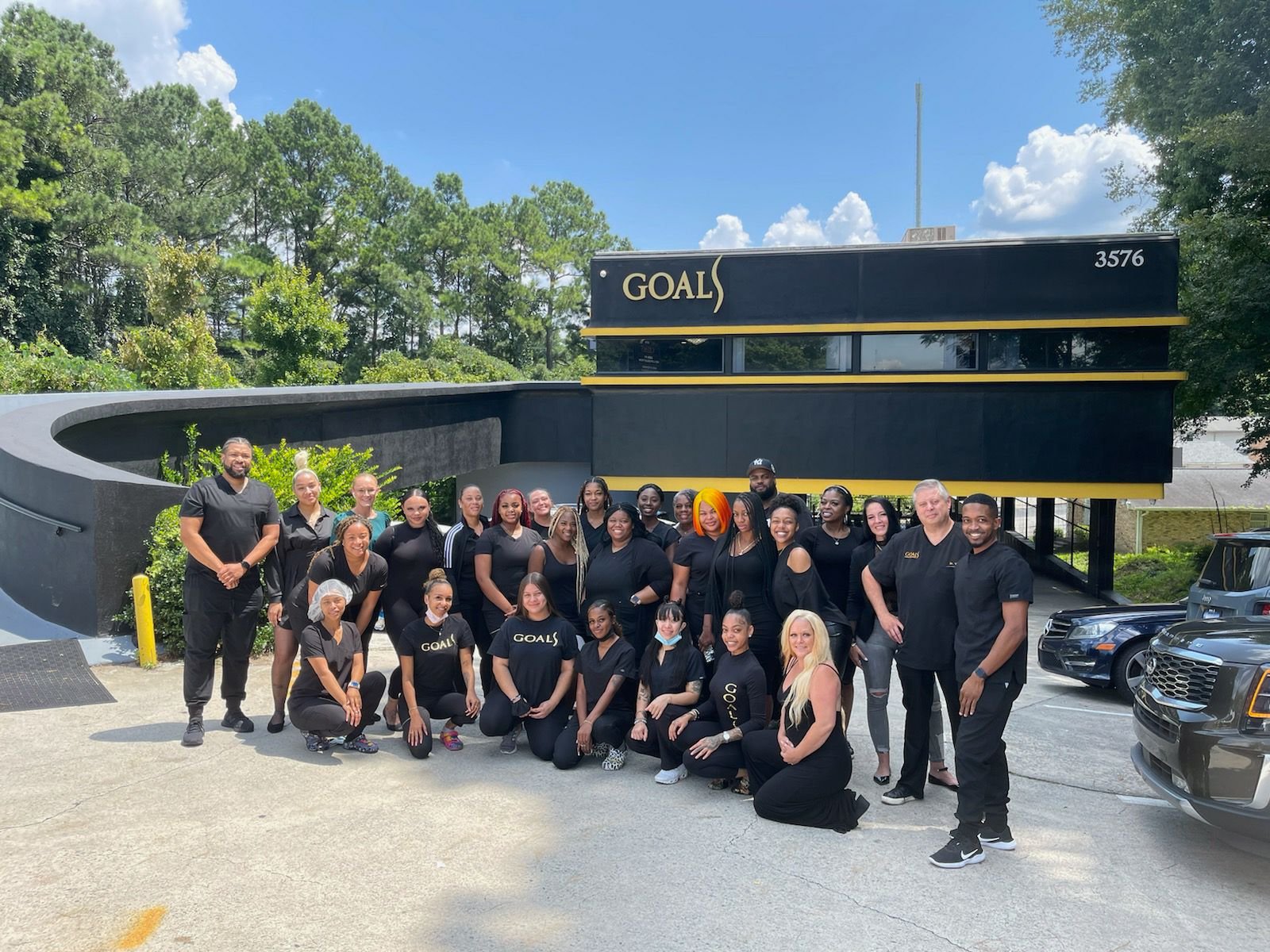 The Goals Aesthetics and Plastic Surgery team, about thirty people dressed in black, outside the practice building with the gold Goals sign