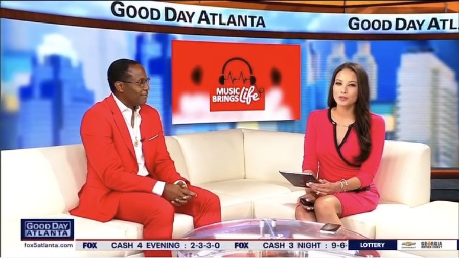 A guest in a red suit on the Good Day Atlanta couch with a host, the Music Brings Life logo on the screen behind them