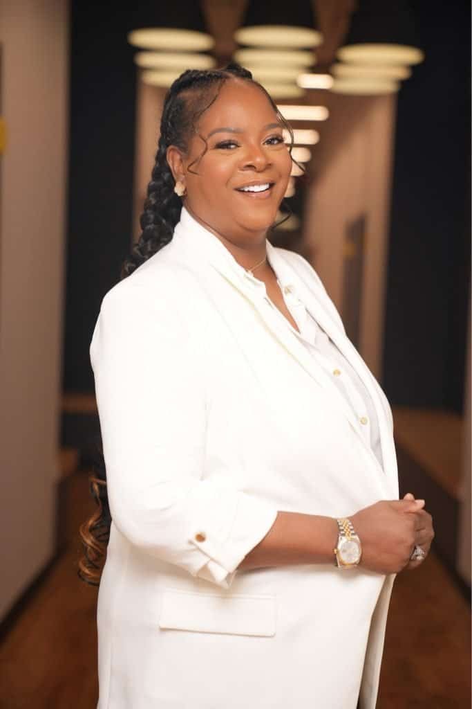 A woman in a white blazer with long braids, smiling in a hallway