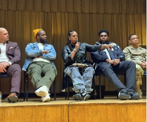 Five speakers seated on a school stage, a woman at the center speaking into a microphone