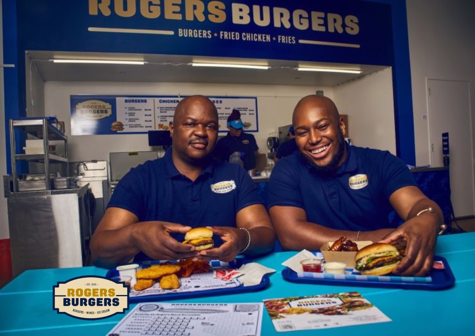 Two men in Rogers Burgers polo shirts at a blue table with burgers and fried chicken, the shop’s counter and menu board behind them
