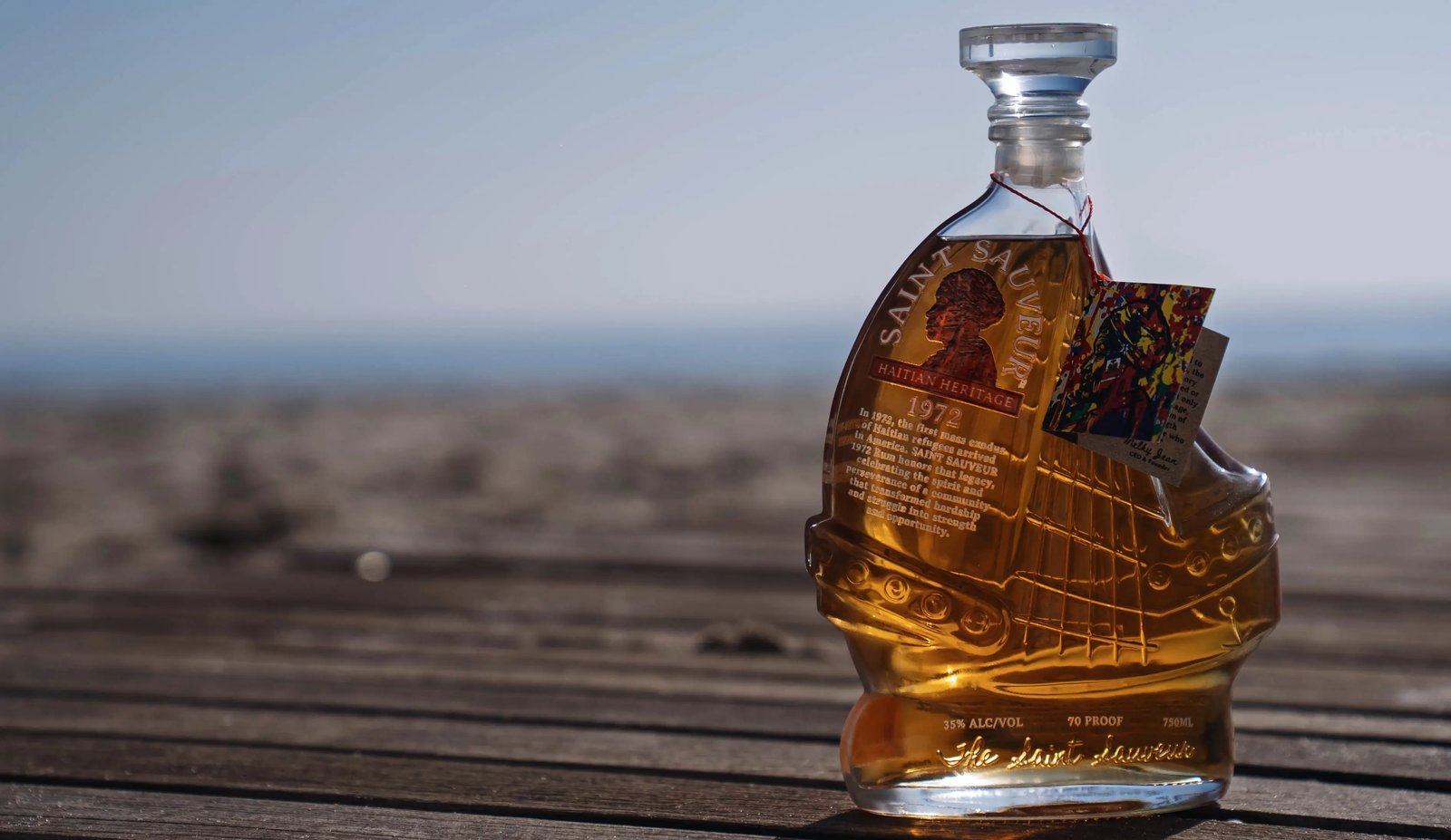 A bottle of Saint Sauveur 1972 rum, shaped like a sailing ship, standing on a wooden boardwalk by the sea