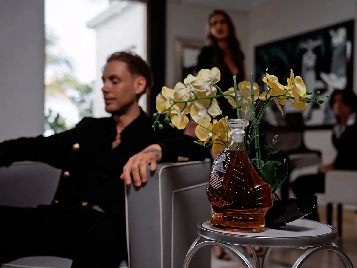 A bottle of Saint Sauveur 1972 beside yellow orchids on a side table, with guests in a living room behind
