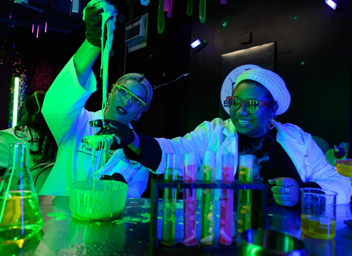 Two guests in lab coats and goggles stretching glowing green slime above a bowl, beside a rack of colored test tubes