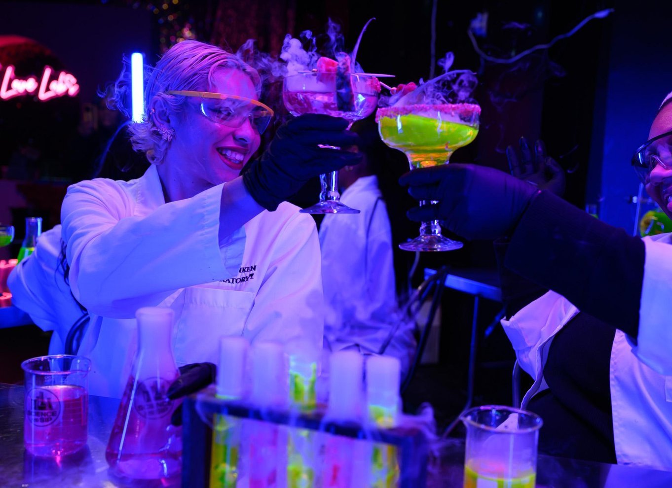 Two guests in lab coats, goggles and black gloves toasting with smoking cocktails in coupe and margarita glasses under ultraviolet light
