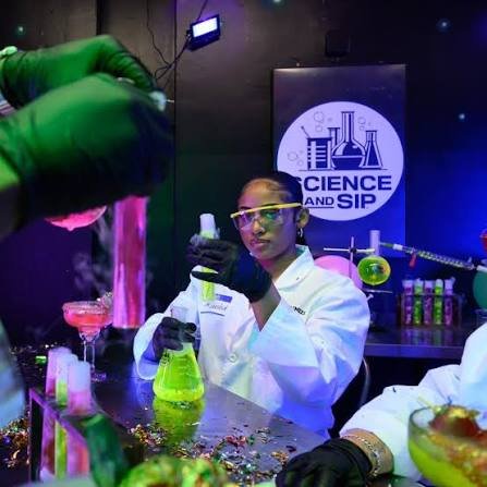 A guest in a lab coat and yellow goggles pouring a glowing green drink from a flask, in front of a Science and Sip sign