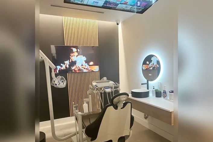 A SmileSpace treatment room with a ceiling-mounted screen, slatted wood feature wall and a backlit round mirror
