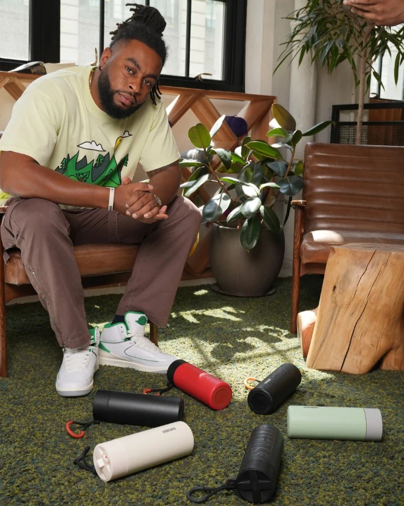 A man seated in a sunlit room, with Suplmnt bottles in several colors lying on the rug around his feet