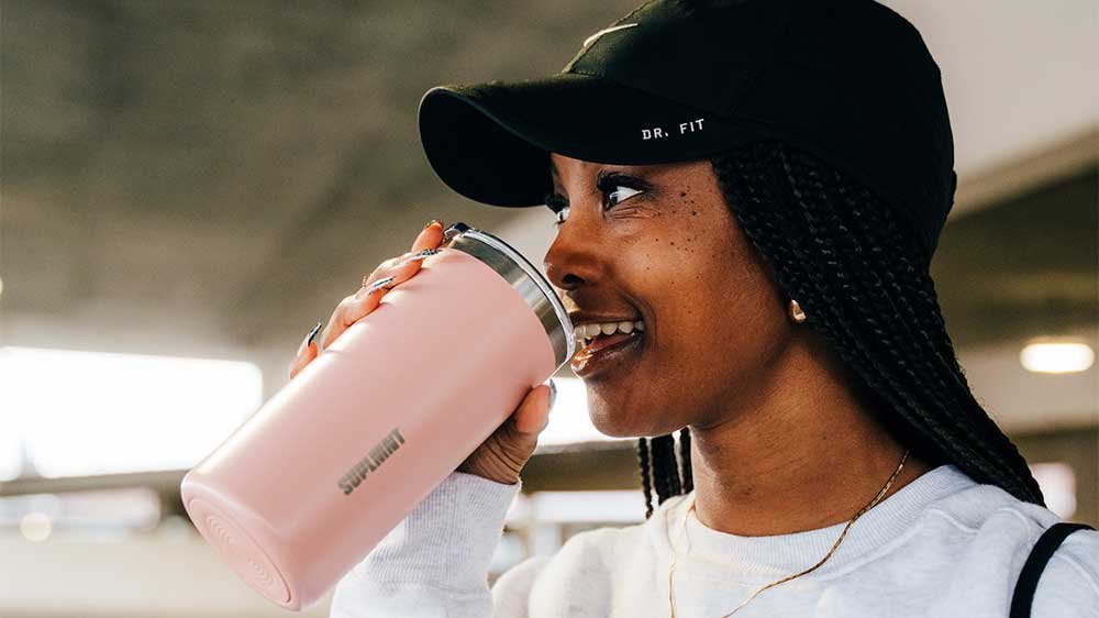 A woman in a black cap drinking from a pink Suplmnt tumbler