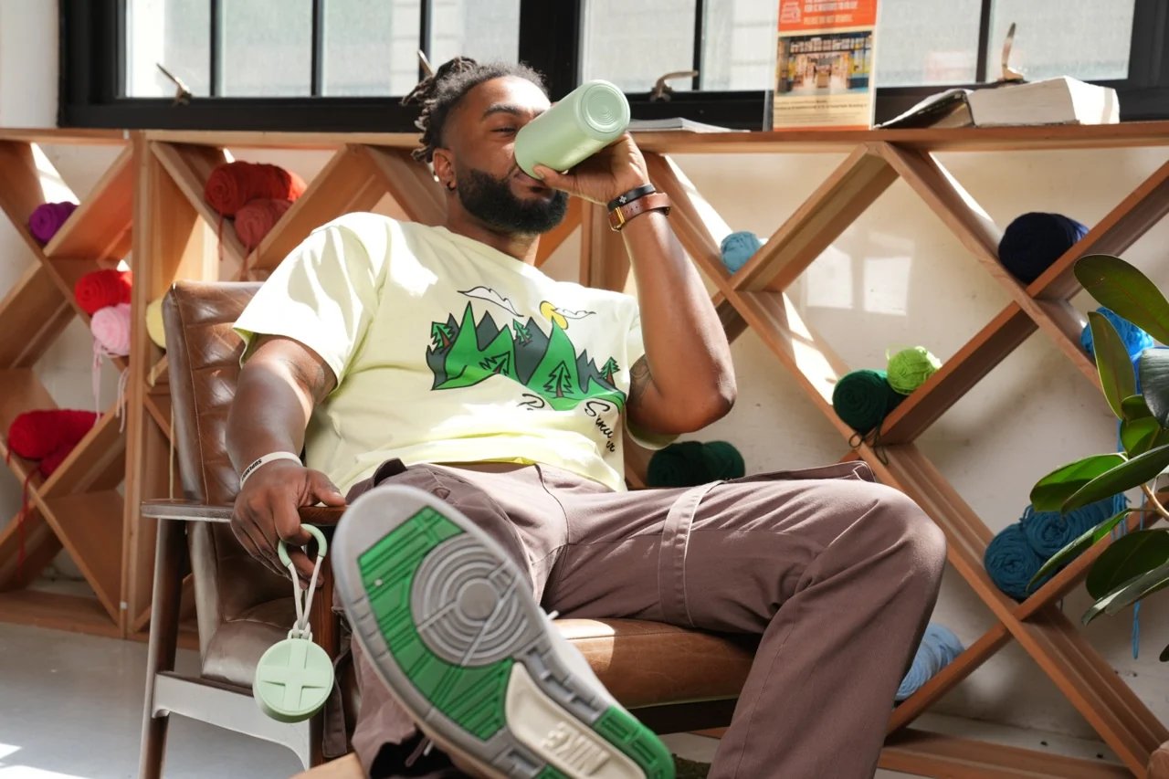 A man in a leather armchair drinking from a mint green Suplmnt bottle, one sneaker raised toward the camera