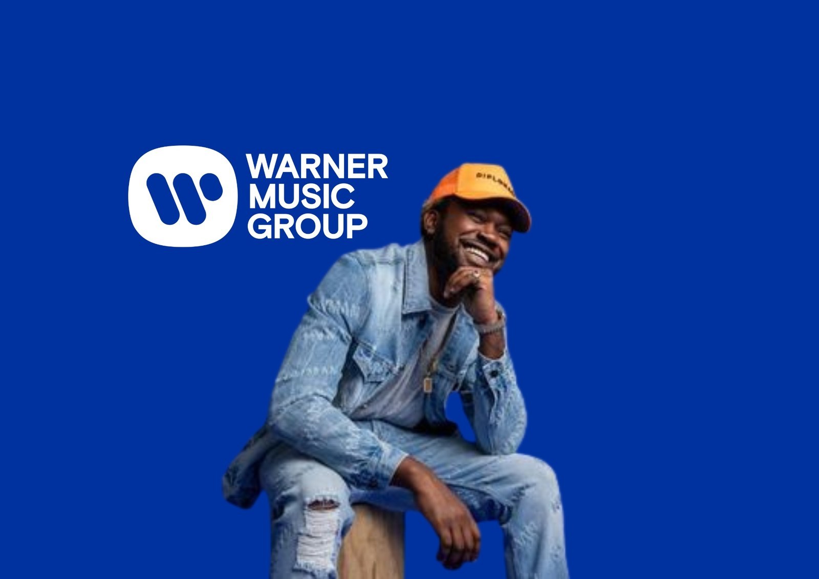 Kranium in a denim jacket and orange cap, seated beside the Warner Music Group logo on a blue ground